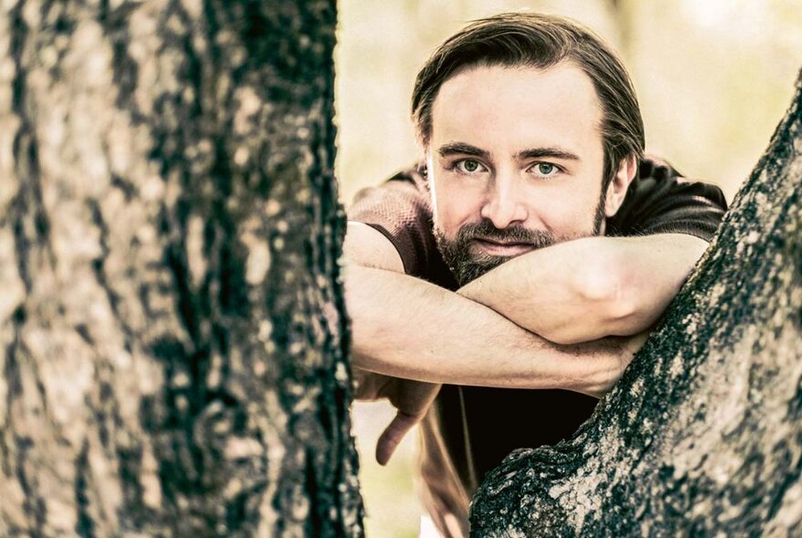 Pianist Daniil Trifonov leaning his chin on his crossed arms through the branches of a tree.