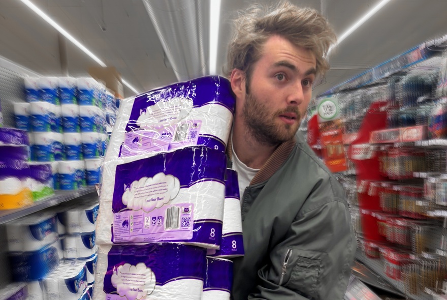 A person in a supermarket aisle carrying multiple packs of toilet tissue in their arms.