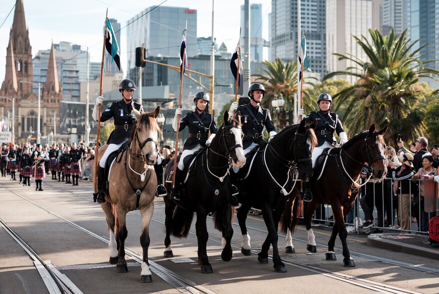 Four mounted servicemen and women marching in front of a crowd at the city skyline of Melbourne.
