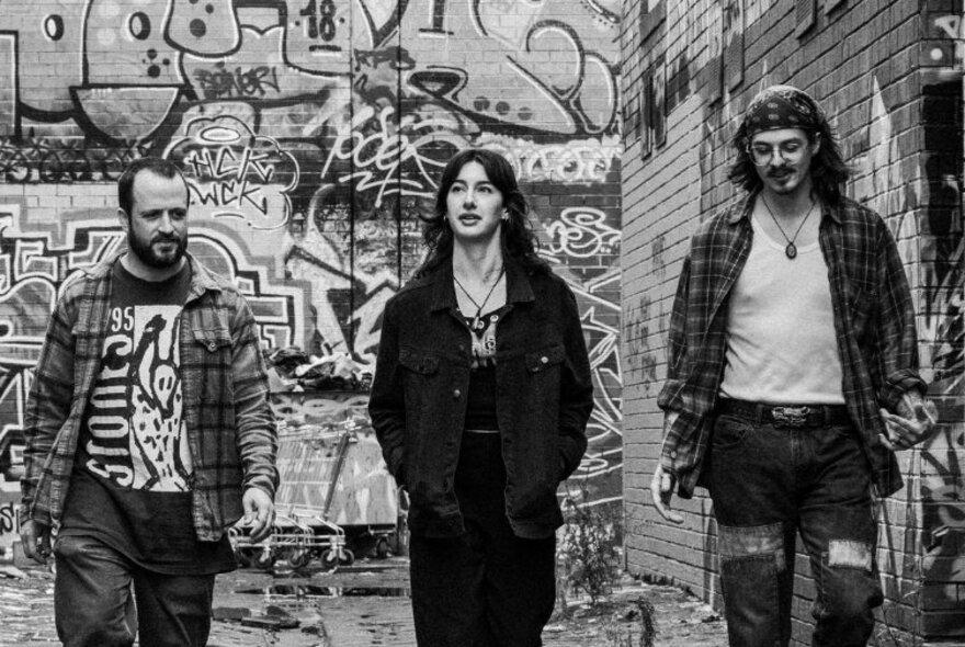 A black and white image of three band members walking along a graffitied laneway. 