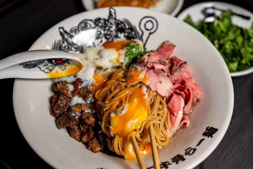 Overhead view of a bowl of ramen, with noodles, sliced meats, egg, with chopsticks and a spoon sticking out of the bowl.