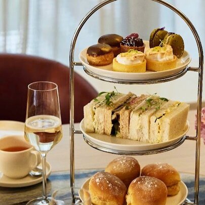 A three-tiered serving platter with scones, sandwiches and profiteroles, alongside a glass of wine, a cup of tea and a small pink and purple floral arrangement.