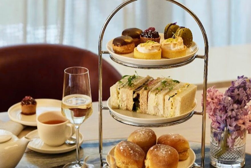 A three-tiered serving platter with scones, sandwiches and profiteroles, alongside a glass of wine, a cup of tea and a small pink and purple floral arrangement.