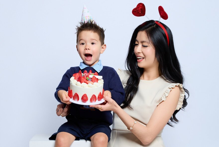 A woman and a young child celebrating a birthday with a strawberry cake, posing for a photo against a white wall.