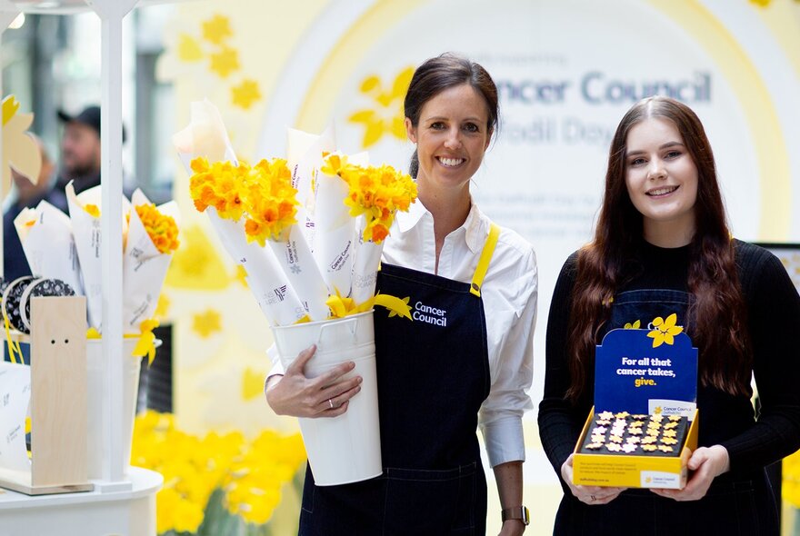 Two smiling people wearing aprons, one holding a bucket of bunches of daffodils, the other a box of daffodil lapel pins.
