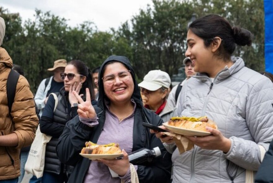 Two people smiling outdoors, enjoying a sausage in bread after picking up litter as part of World Cleanup Day, other people in the background.