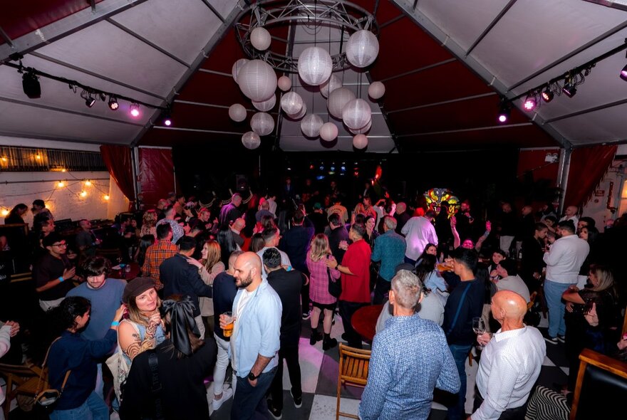 People gathered under a tented outdoor event roof with hanging lights.