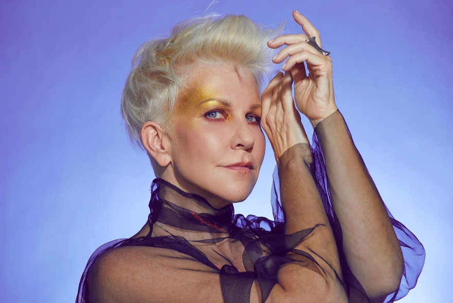 Joyce DiDonato in a sheer black top with her hands raised towards her head against a violet background. 