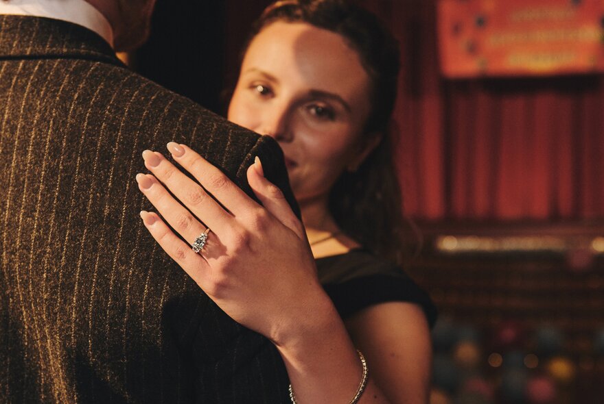 A woman's left hand on a man's shoulder, highlighting the engagement ring on her finger.