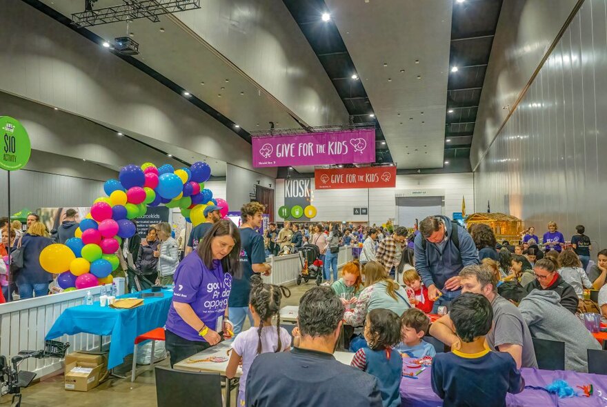 Children and adults seated at rows of tables engaged in activities in a large room with banners and balloons.