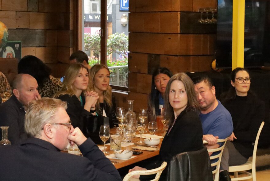 People at a table on a restaurant with many wine glasses in front of them, listening to a speaker.