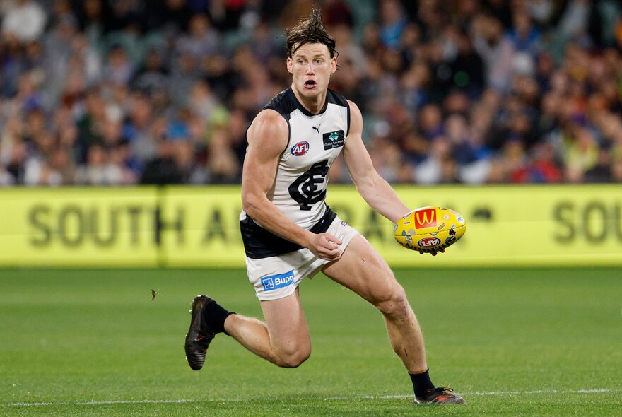 A Carlton AFL football player running with the ball in a stadium during a footy match.