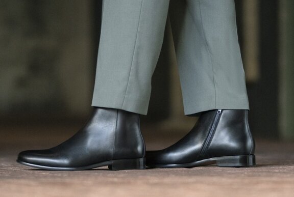 A pair of well-made black leather boots, being worn by a person in grey trousers.