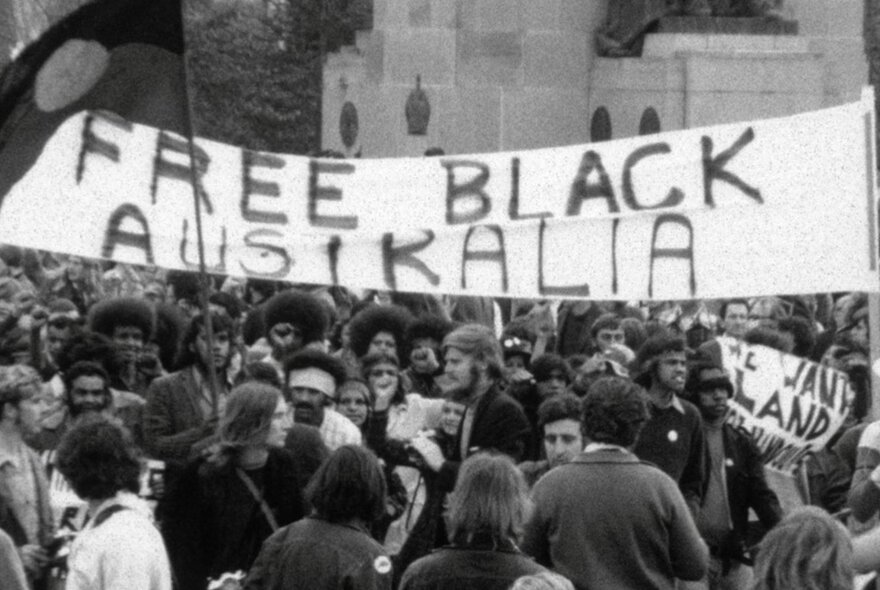 Archival photo and still from a documentary features Indigenous rights activists and supporters marching under a 'Free Black Australia' banner during the historic 1972 protests connected to the foundation of the Aboriginal Tent Embassy.