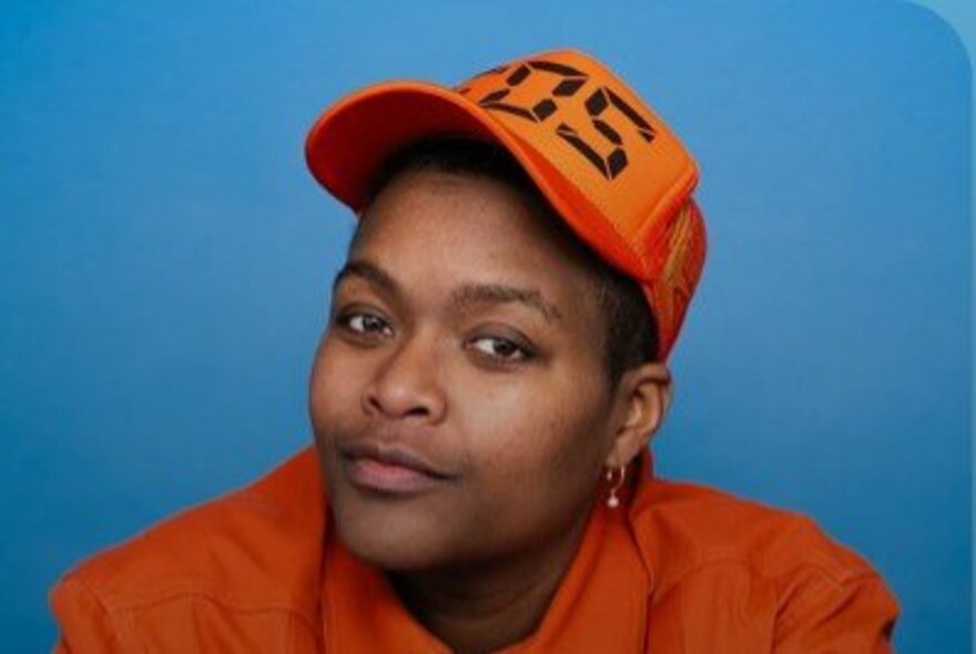 Portrait shot of American comedian, Sam Jay, wearing an orange trucker cap and orange jacket; blue background.