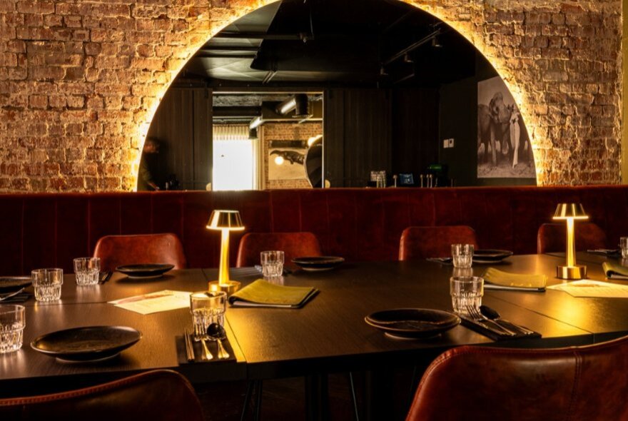Moodily lit restaurant interior with semicircular backlit mirror, brick wall and lamplit tables laid for dinner.