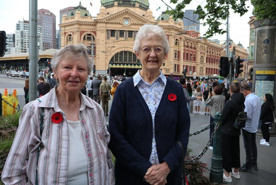 Two older women wearing Remembrance Day poppies in the city with Flinders Street Station in the background. 