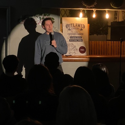 Comedian performing on a small stage, spotlit in front of seated audience.