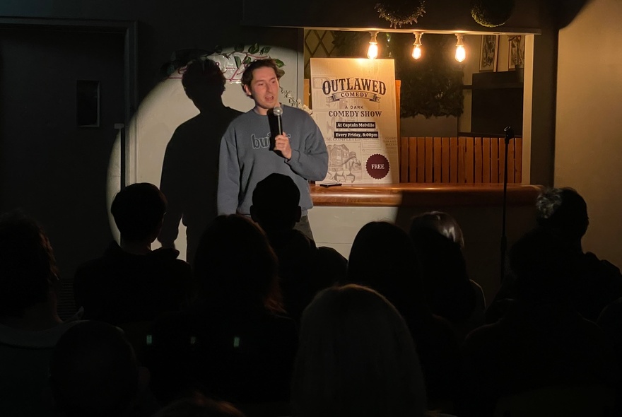 Comedian performing on a small stage, spotlit in front of seated audience.