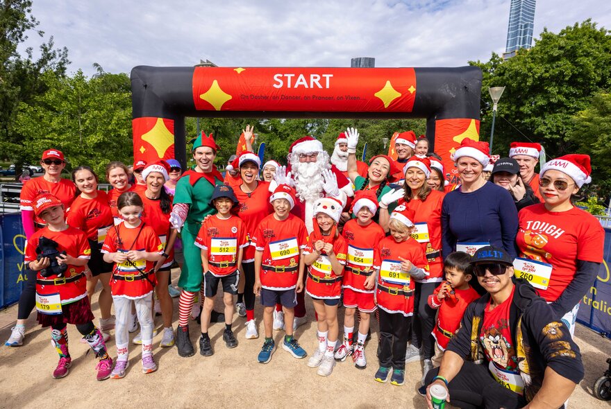 Large group of people wearing Santa hats next to Santa under the start line of a race.