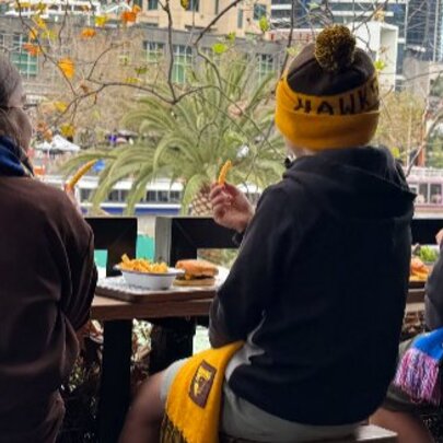 Children seated overlooking the Yarra, viewed from behind, wearing footy hats and scarves