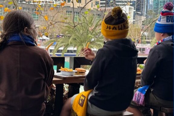 Children seated overlooking the Yarra, viewed from behind, wearing footy hats and scarves