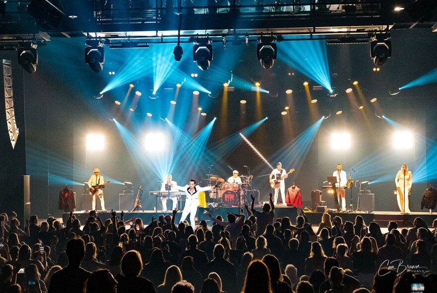 A band performing on a stage in front of a crowd in a large venue, with theatrical lighting.