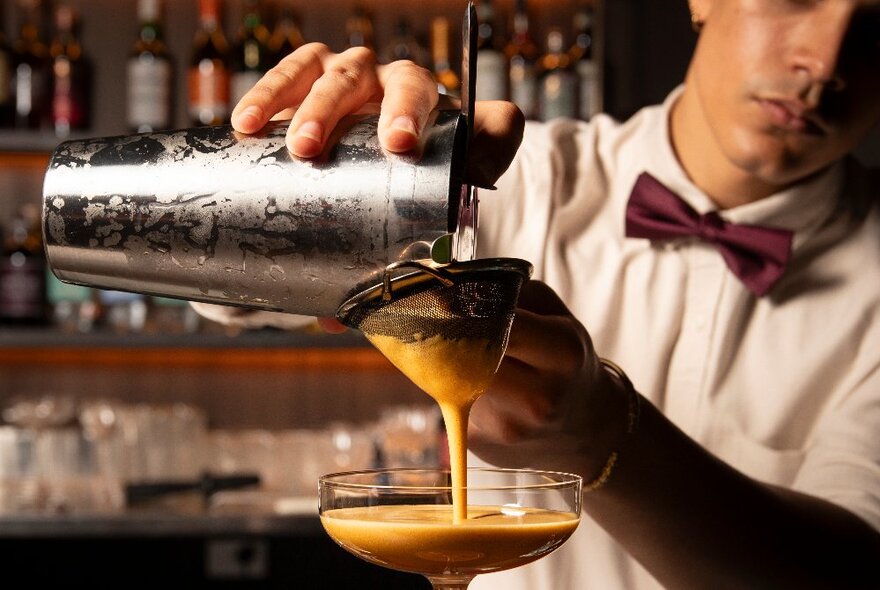 A bow-tied bartender pours a frothy cocktail from a shaker through a sieve.