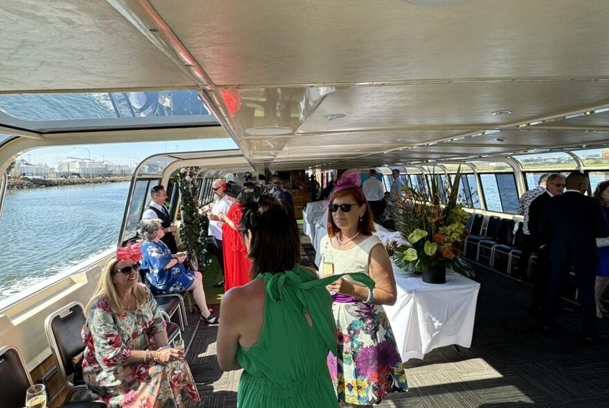 A group of racegoers on board a boat, all dressed in formal racing wear and hats, enjoying food and drinks.