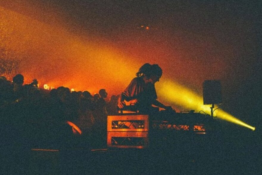 A DJ in silhouette at a mixing desk performing in a darkened space with people in the background dancing.