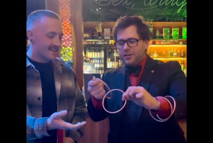 A magician holding two joined rings in a pub setting with another man outstretching his hand to take them. 