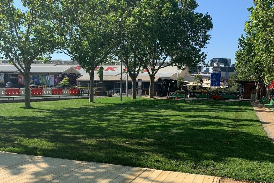 Green expanse of lawn with trees providing leafy shade and the Queen Victoria Market sheds visible in the background.