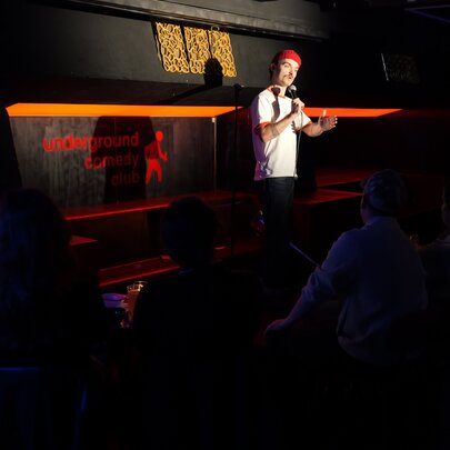 A live stand-up comedy performance in a dimly lit basement venue or club with an audience seated in the foreground.