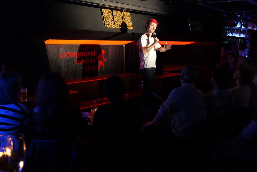 A live stand-up comedy performance in a dimly lit basement venue or club with an audience seated in the foreground.