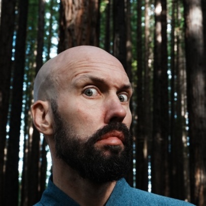 Bald man with beard in a forest setting, looking shocked.