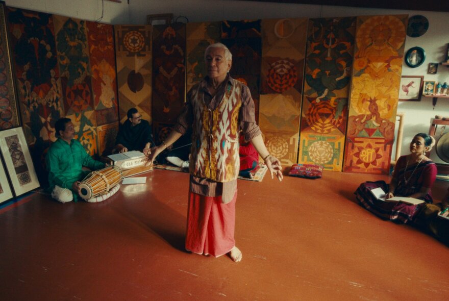 An older Indian man dancing in a sunlit room with colourful Indian motifs on the wall behind him.