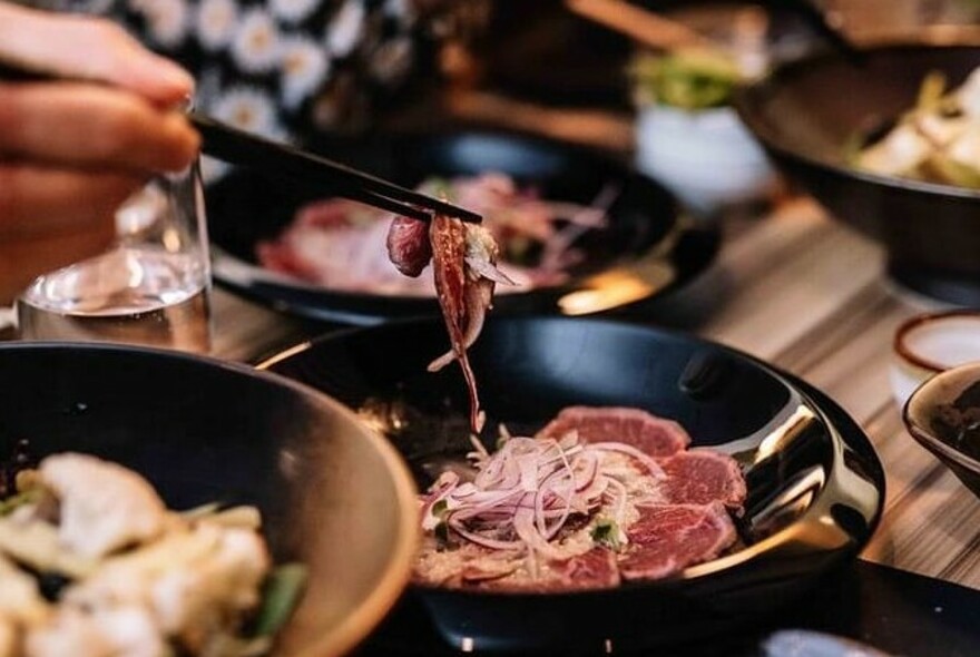 Several plates of food and a person's hands using chopsticks. 