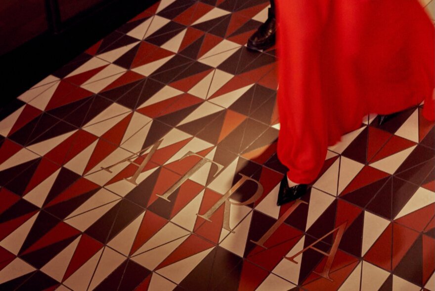 A striking tiled floor with the word Atria in gold and the red edge of a dress moving across the space.