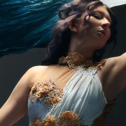 A person wearing an ethereal, one-shoulder white gown adorned with intricate golden floral appliques and a matching textured collar.