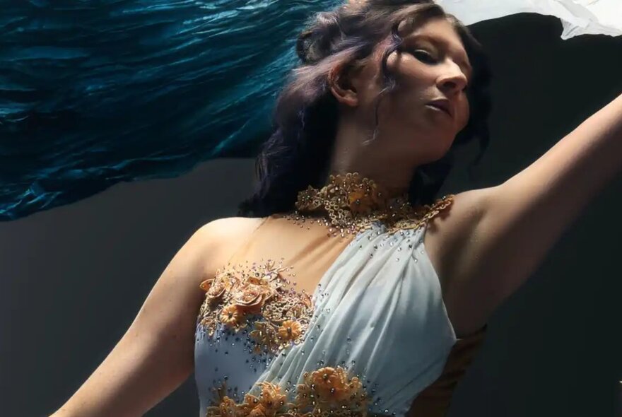 A person wearing an ethereal, one-shoulder white gown adorned with intricate golden floral appliques and a matching textured collar.