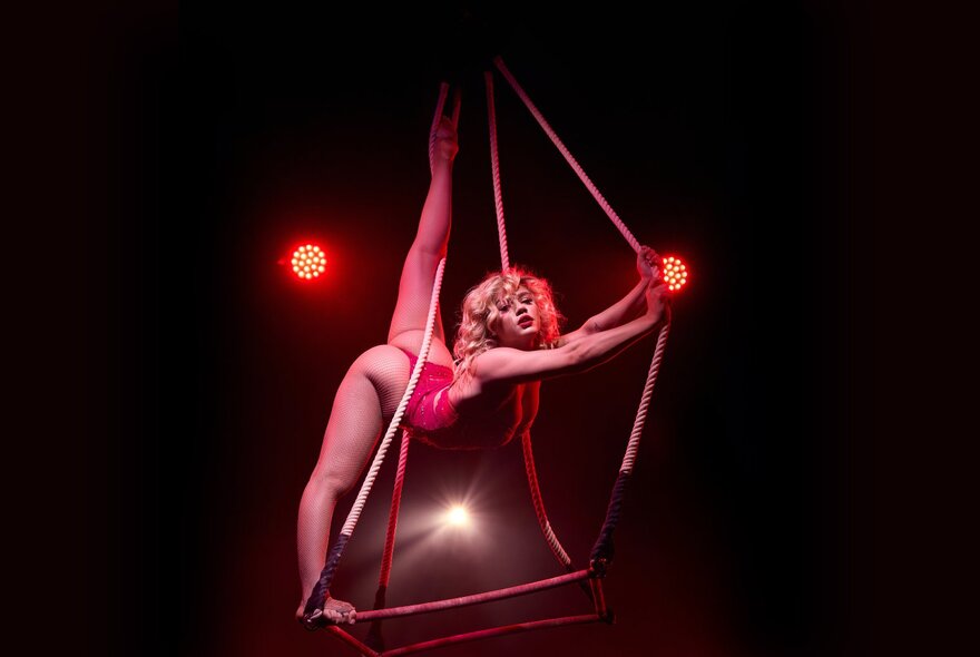 A circus performer in a red burlesque costume doing the splits on a rope trapeze.