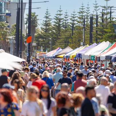 A busy outdoor market with colourful tents lining a pedestrian walkway alongside tall palm trees and crowds of visitors.