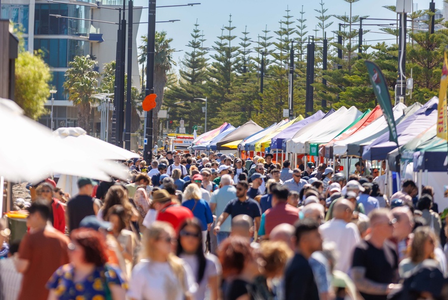 A busy outdoor market with colourful tents lining a pedestrian walkway alongside tall palm trees and crowds of visitors.