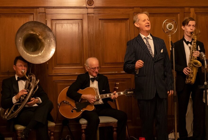 A swing band playing live music in front of a wood panelled wall, with a man singing into an old-fashioned style radio microphone.