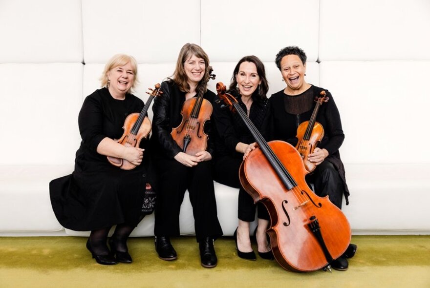 Four members of a string quartet all sitting in a bench with their instruments smiling warmly. 