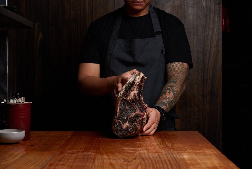 A chef in a black apron holding up a large dry-aged steak