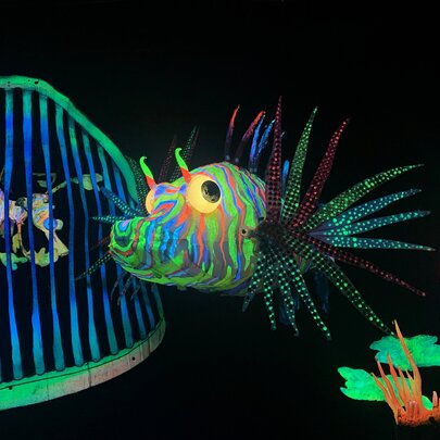 A blacklit underwater scene showing a colourful lion fish and a leafy sea dragon behind a large set of teeth.