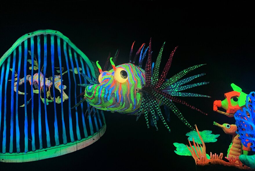 A blacklit underwater scene showing a colourful lion fish and a leafy sea dragon behind a large set of teeth.