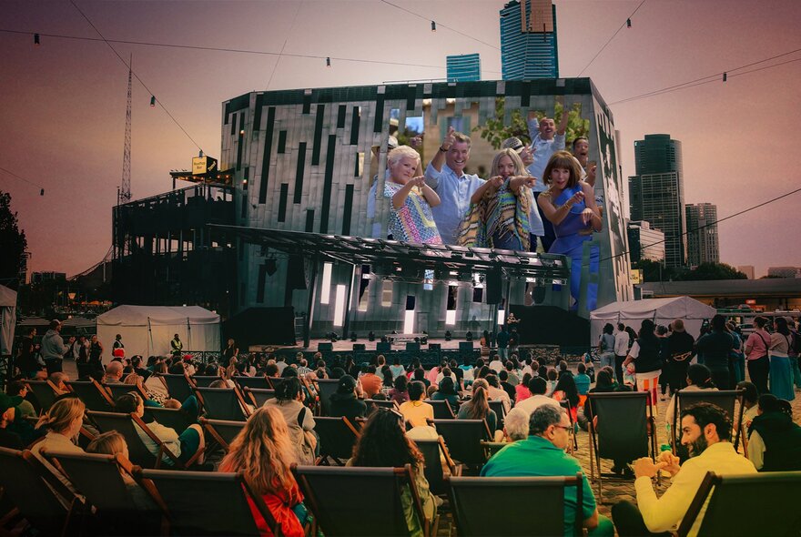 People seated in Federation Square watching a film on the Big Screen, a pinky dusk glow in the sky. 