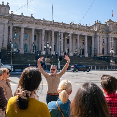 The back of a group of people on a walking tour in front of a group leader with his hands in the air at Parliament House. 